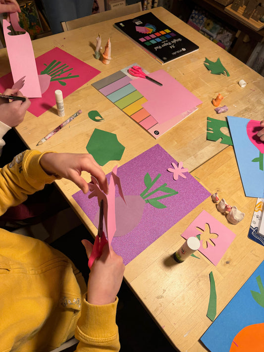 A child's hands and arms in a yellow sweatshirt are shown cutting pink paper with scissors at a wooden craft table surrounded by colorful paper cutouts, glue, and art supplies.