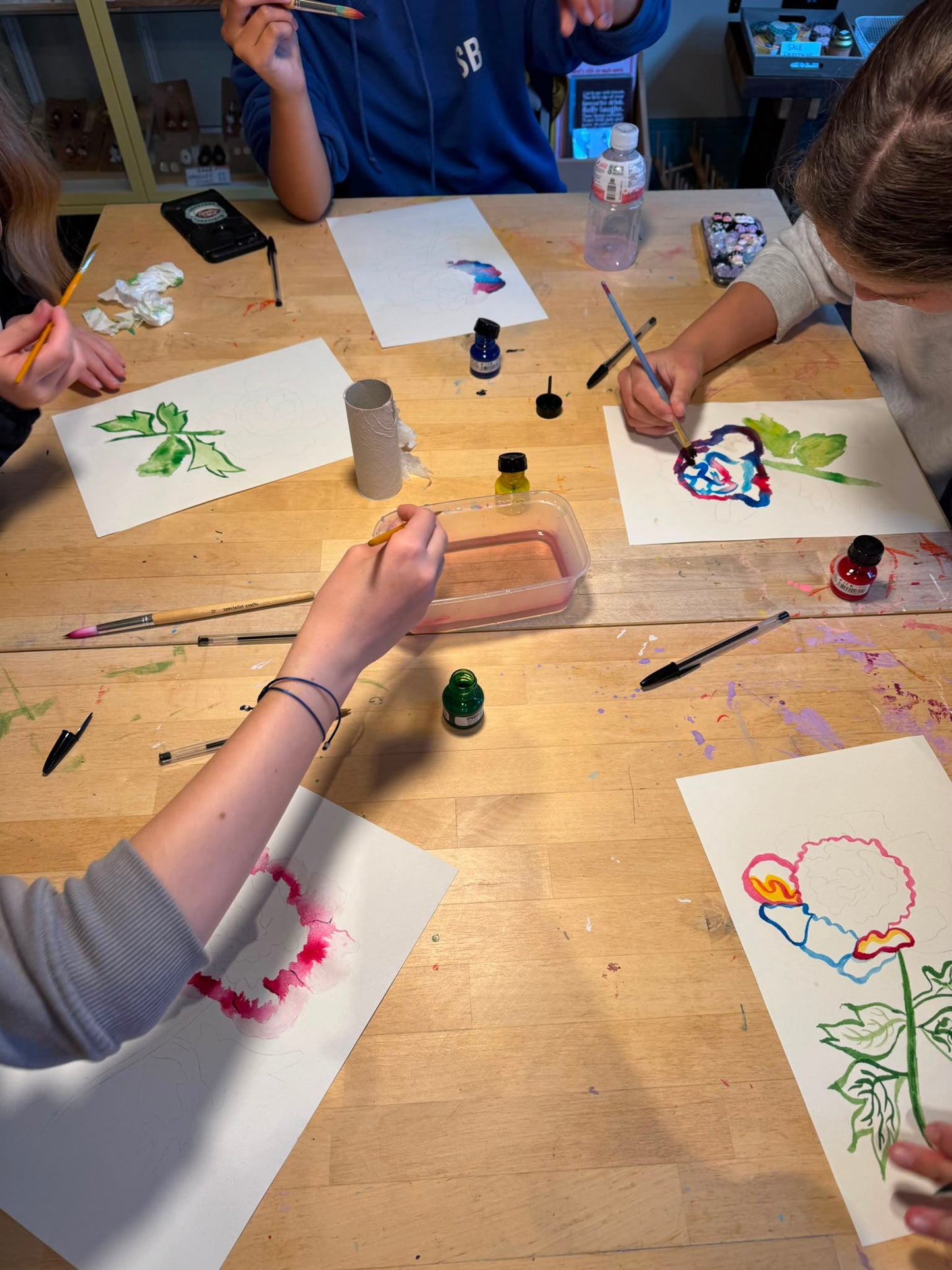 A group of children painting on white paper sheets spread across a light wood table, using small bottles of paint and brushes.