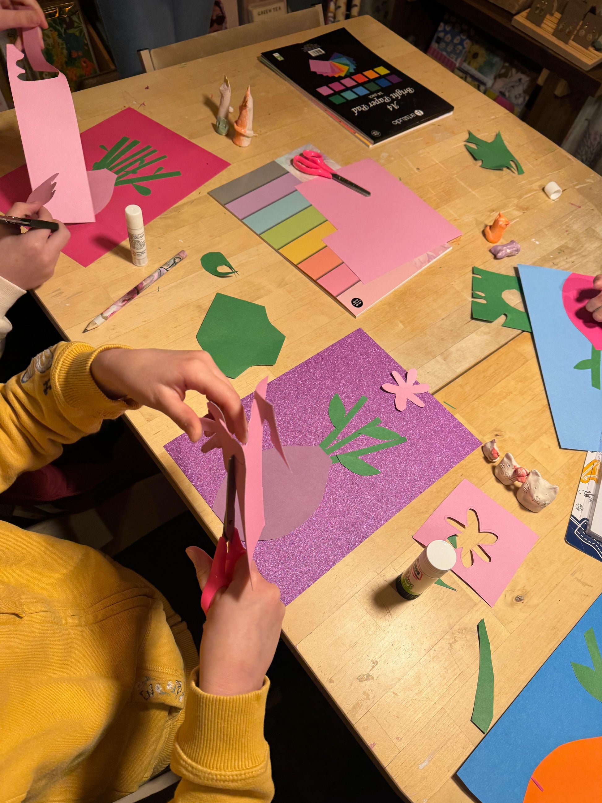 A child's hands and arms in a yellow sweatshirt are shown cutting pink paper with scissors at a wooden craft table surrounded by colorful paper cutouts, glue, and art supplies.