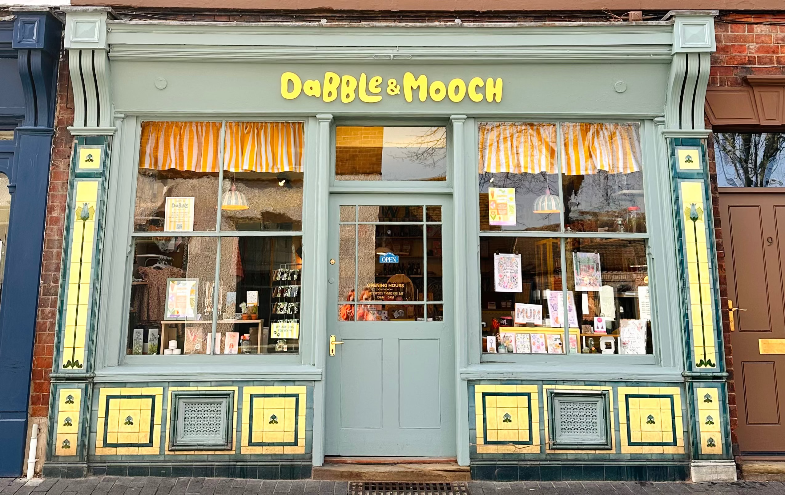 A storefront facade of a brick building with a pale green shop front reading 'DaBBLE & MooCH' in yellow letters. The shop has large windows, striped orange-and-white curtains, and decorative yellow-and-green trim.
