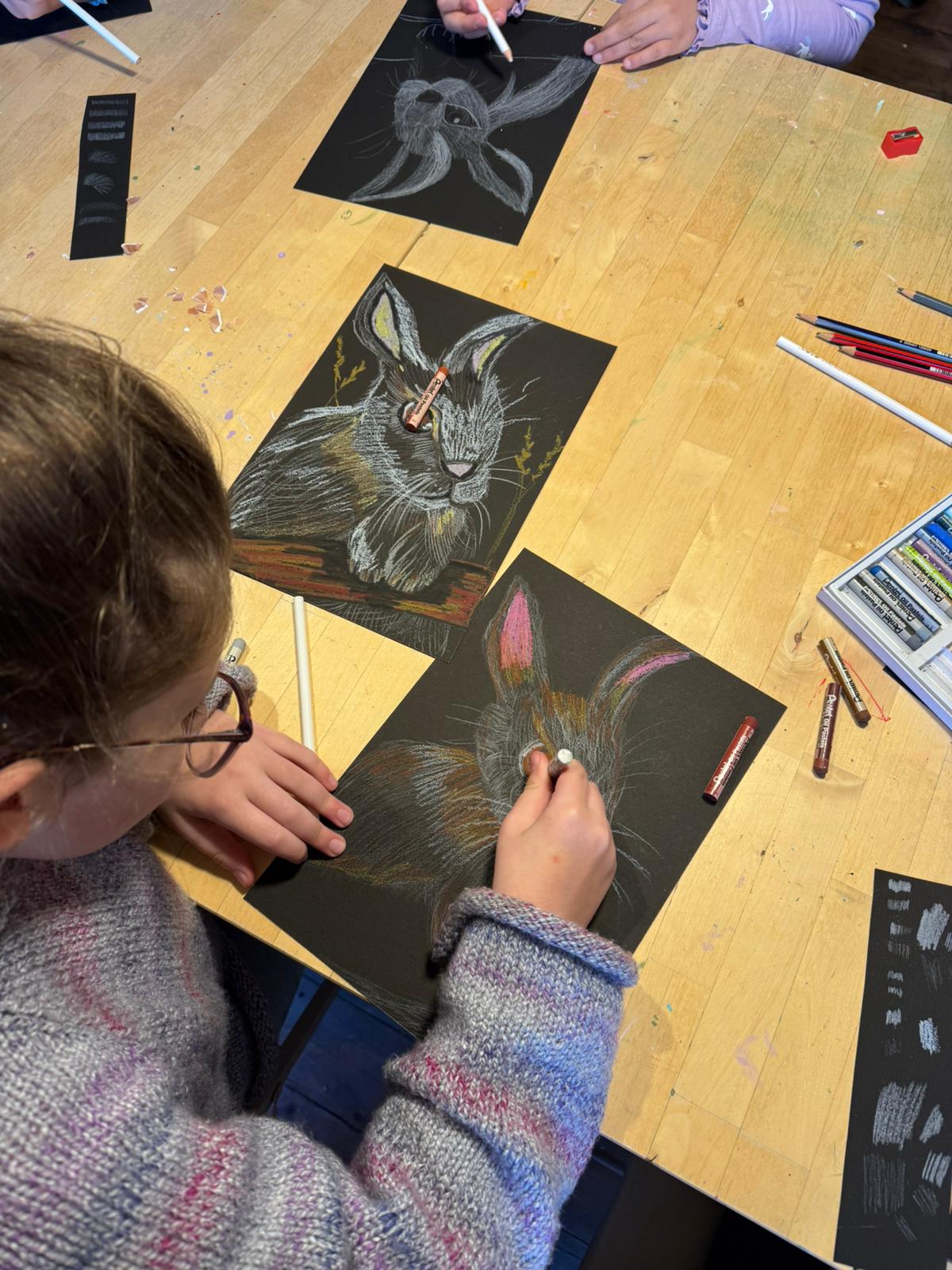 A child drawing a rabbit on a black sheet of paper at a wooden table, with other drawings and colored pencils visible around the workspace.
