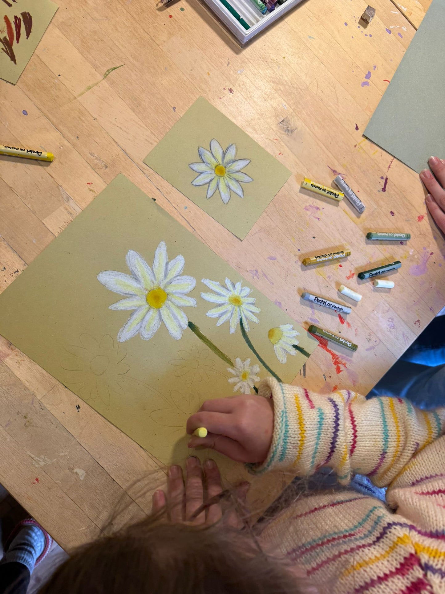 A child's hand drawing white daisies with yellow centers on tan paper using a yellow crayon, set on a wooden craft table surrounded by crayons.