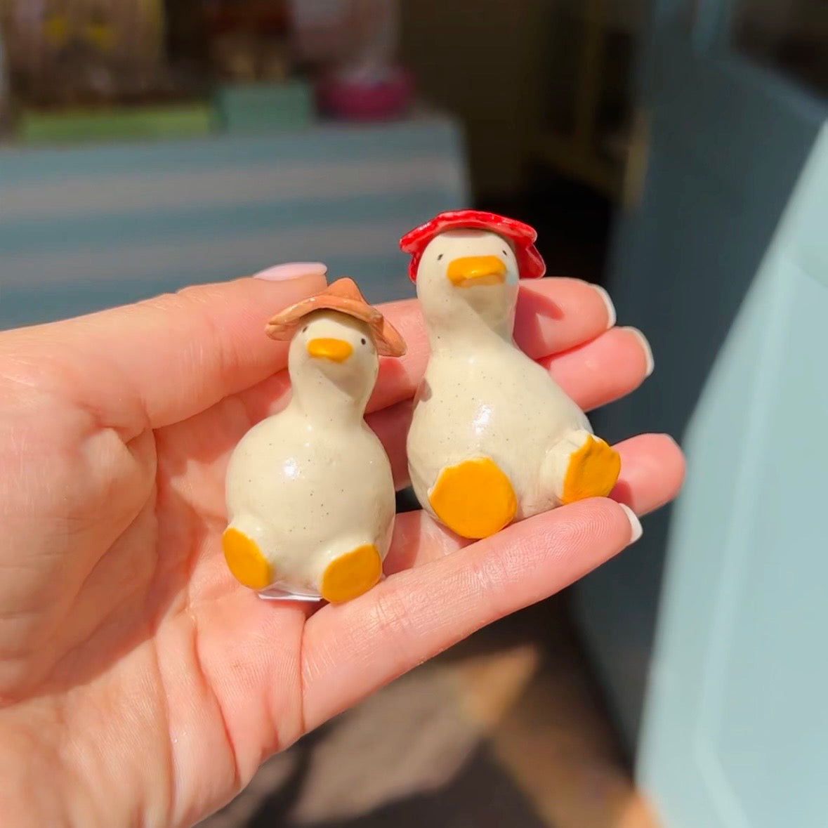 Two small duck figurines held in a person's hand. One duck wears a tan hat and the other wears a red hat, both with cream bodies, orange beaks, and yellow feet.