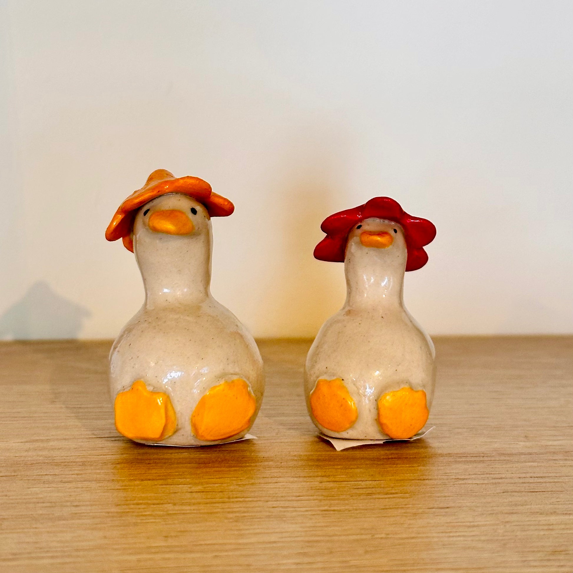 Two small ceramic duck figurines sitting side by side on a wooden surface against a plain light background. The left duck wears an orange hat and has yellow feet, while the right duck wears a red hat and has yellow feet.