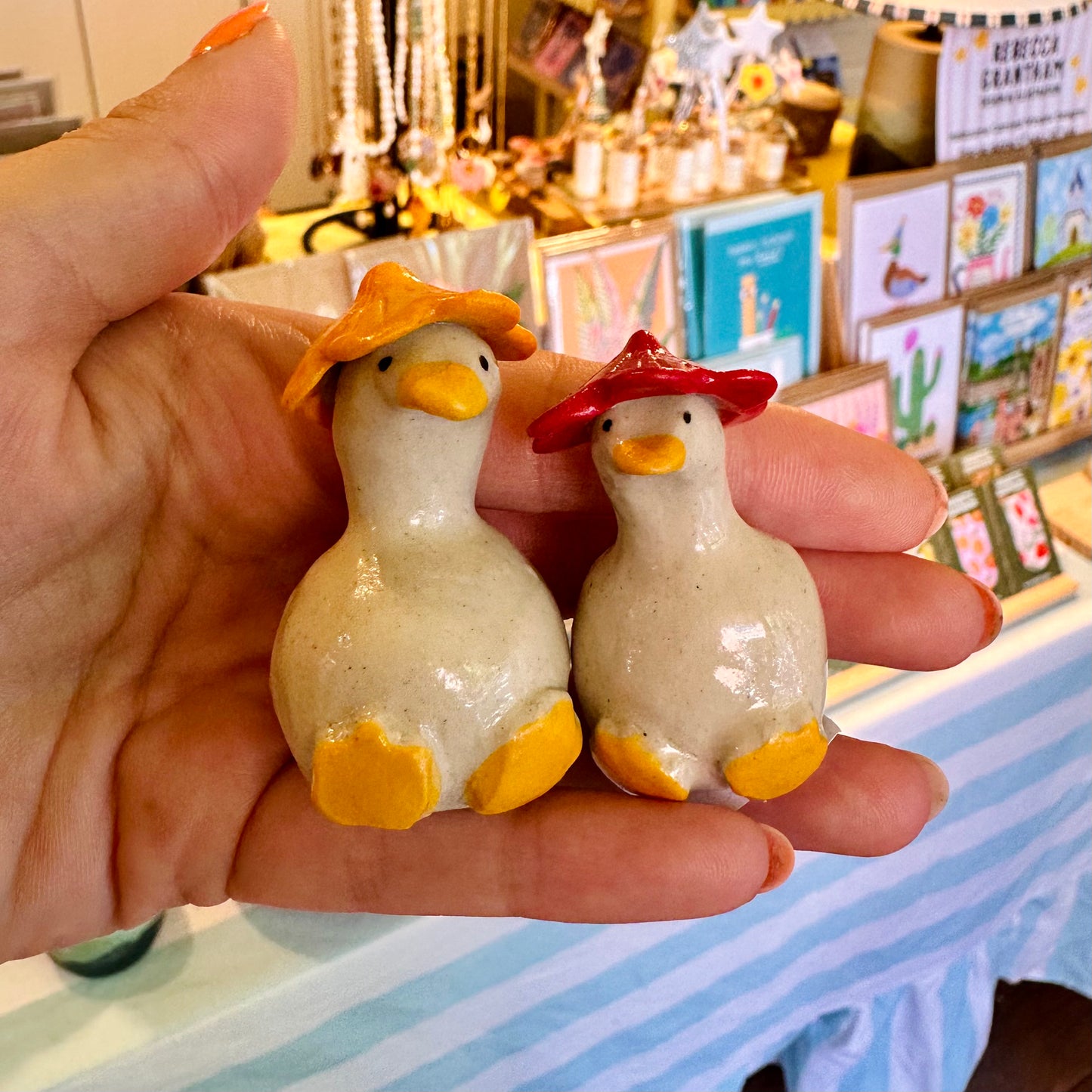 Two small ceramic duck figurines with yellow beaks and feet, one wearing a yellow hat and the other wearing a red hat, held in a person's hand in a shop setting.