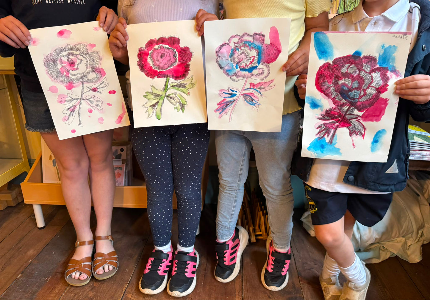 Four children standing together and holding up four different watercolor flower paintings on paper, showing colorful floral designs in red, pink, blue, and green.