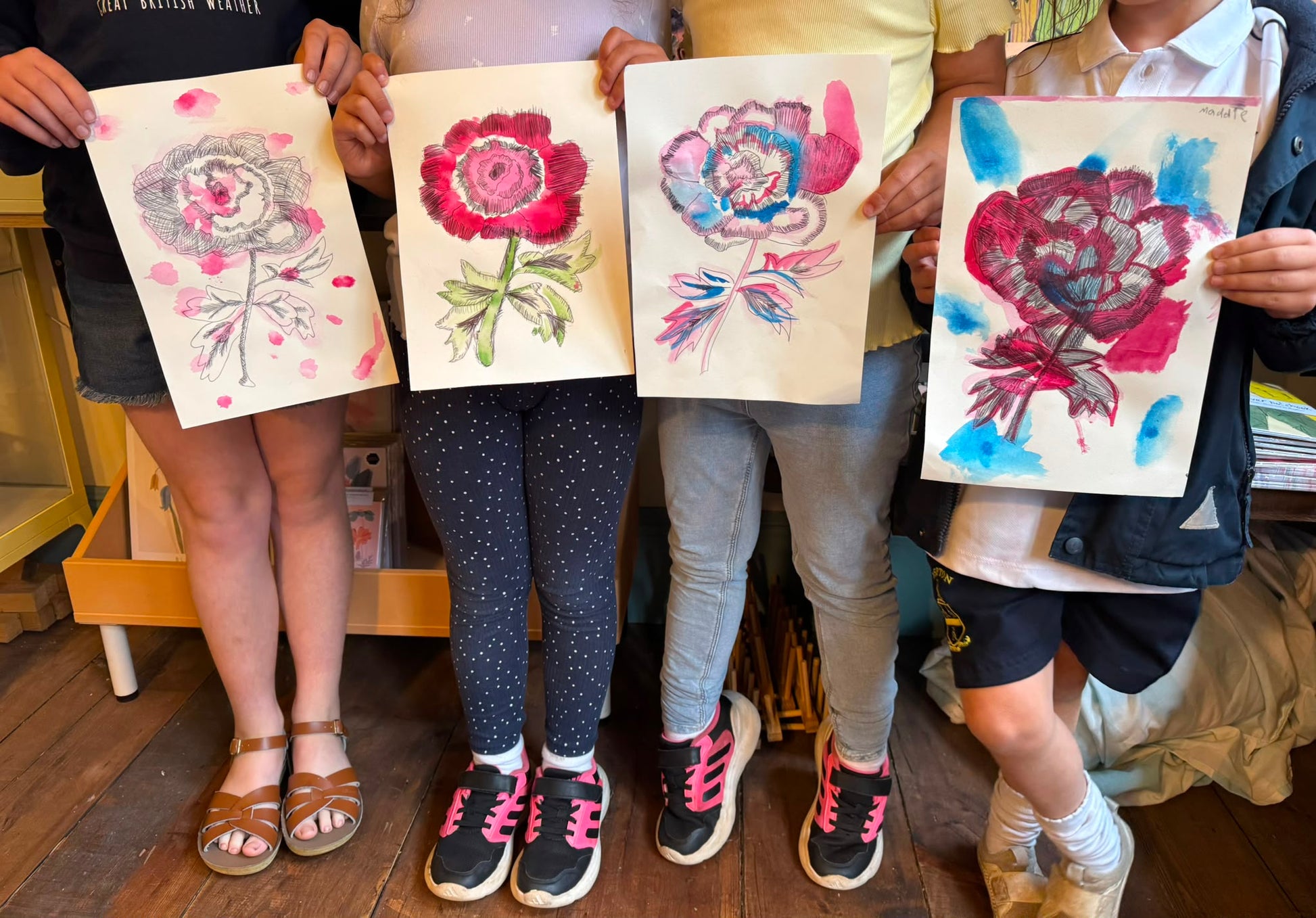 Four children standing together and holding up four different watercolor flower paintings on paper, showing colorful floral designs in red, pink, blue, and green.