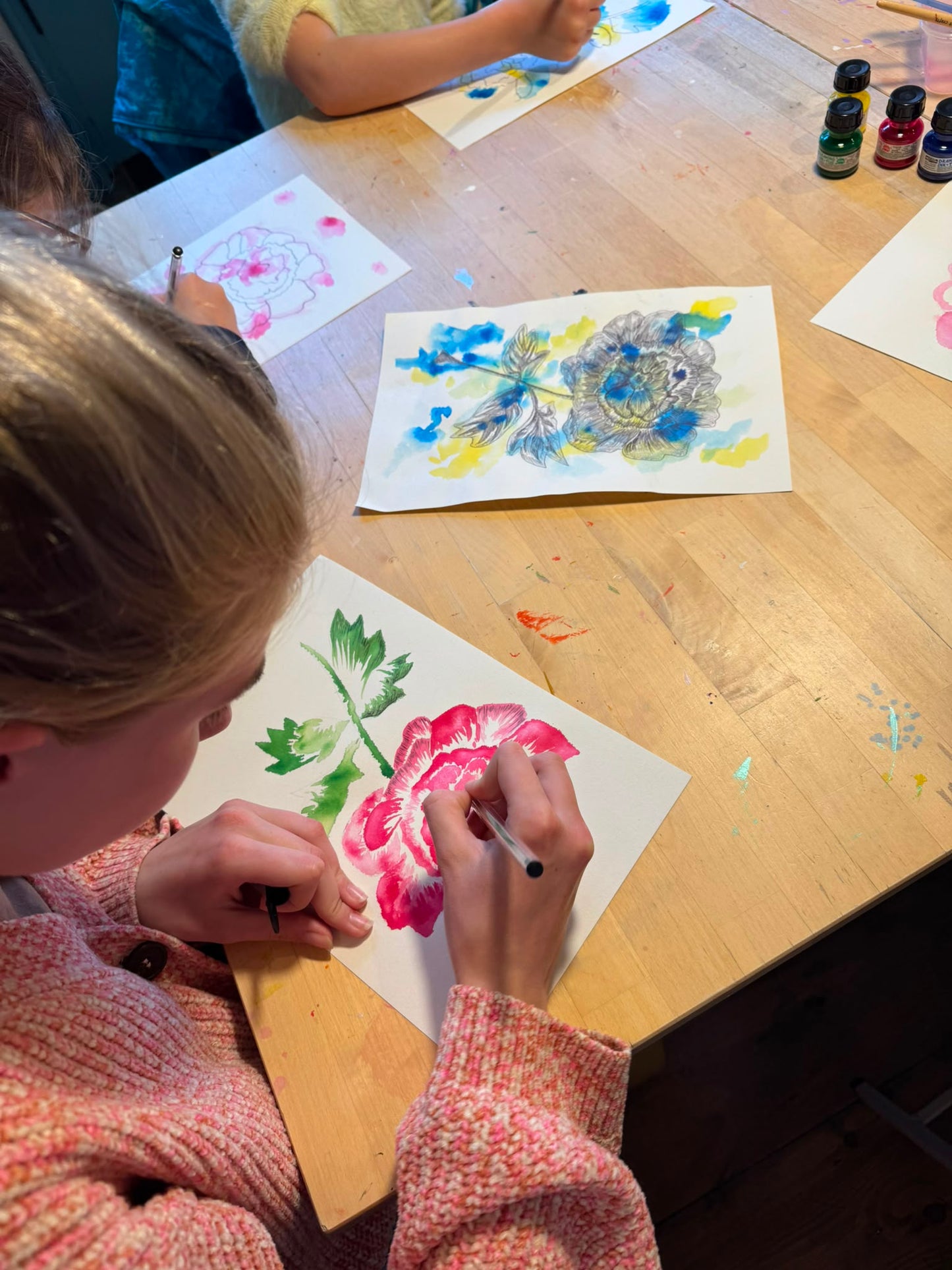 A child painting a pink flower on white paper at a wooden table, with other colorful artwork and paint bottles visible in the scene.