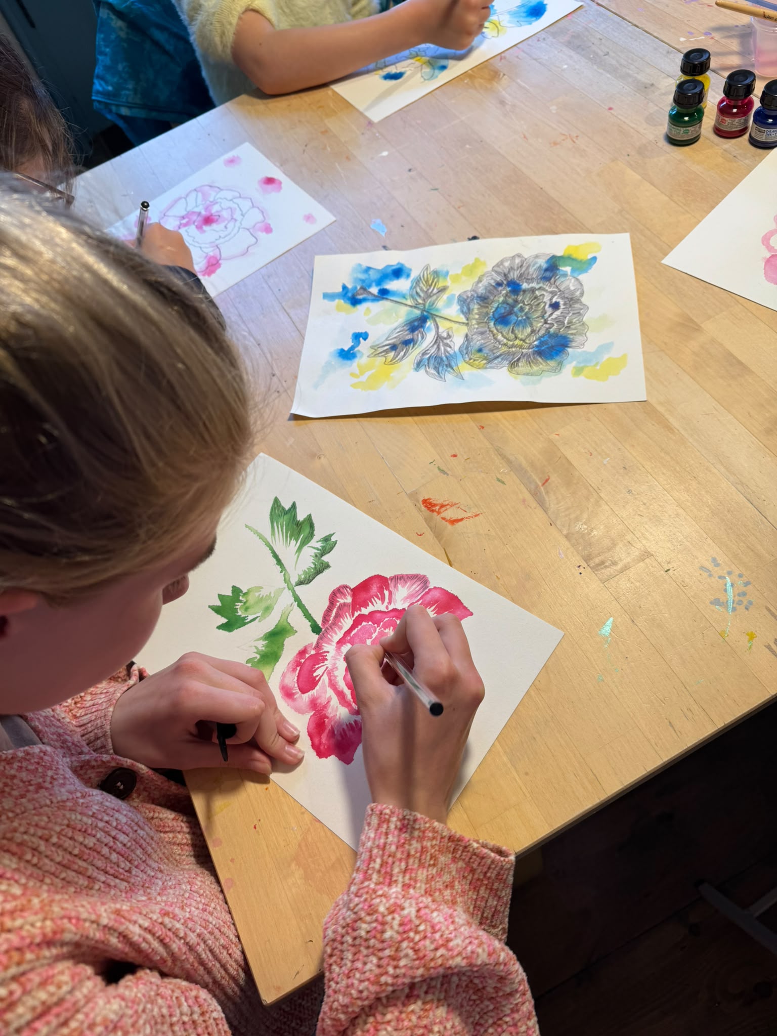 A child painting a pink flower on white paper at a wooden table, with other colorful artwork and paint bottles visible in the scene.