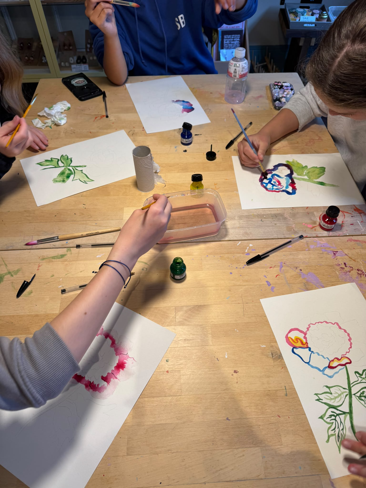 A group of children painting on white paper sheets spread across a light wood table, using small bottles of paint and brushes.