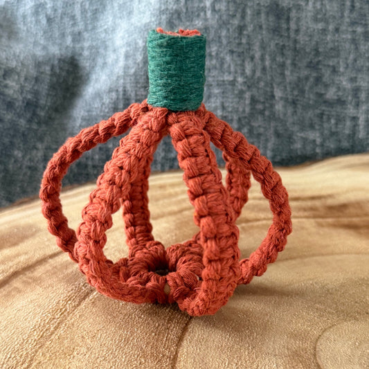 A small crocheted pumpkin decoration with a rust-orange body and a dark green stem, displayed on a wooden surface against a gray fabric backdrop.