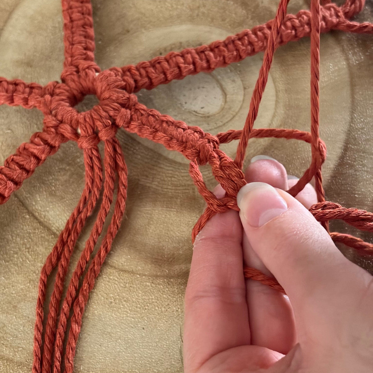 A close-up of a hand tying a knot in a terracotta red macrame cord, set against a natural wood background.